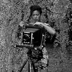 Deana Lawson stands behind a large camera in a wooded area.